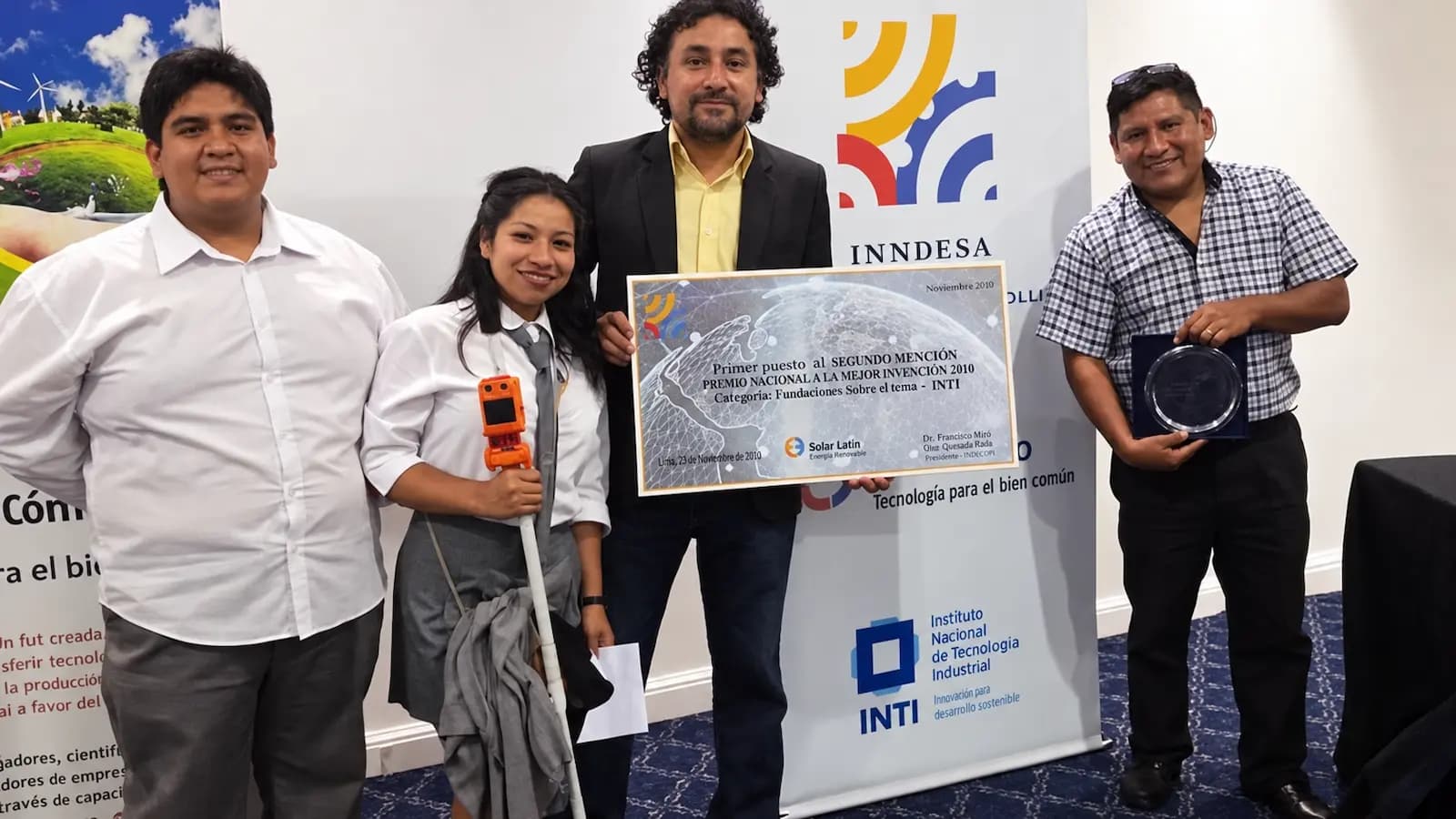 Alumnos y docentes con el bastón inteligente y el diploma del premio INNDESA, frente a los banners del INTI